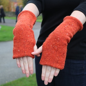 May include: A pair of orange arm warmers with a textured knit design. The warmers are a vibrant orange color with flecks of white throughout. The cuffs are ribbed, and the warmers are shown on a person's arms.