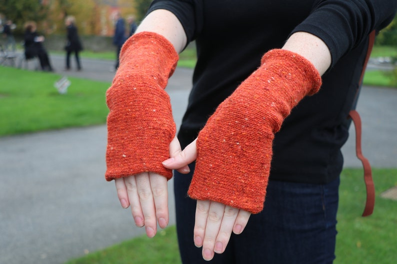 May include: A pair of orange arm warmers with a textured knit design. The warmers are a vibrant orange color with flecks of white throughout. The cuffs are ribbed, and the warmers are shown on a person's arms.