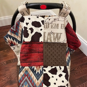 May include: A patchwork car seat cover with a red, brown, white, and blue color scheme. The cover features a cow print, a red and white striped pattern, and a geometric pattern. The text "Ride 'em Up Law" is printed on the cover.