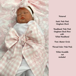 May include: A baby swaddled in a pale pink and white gingham check sash. The baby is wearing a matching headband with a white headband. The sash is embroidered with a monogram in pale pink thread. The text on the right side of the image reads "Pictured".