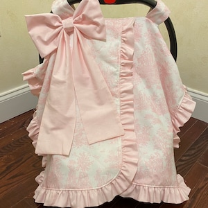 May include: Pink car seat cover with a large pink bow and a white and pink floral pattern. The cover has ruffles around the edges.