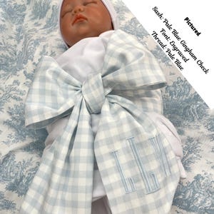 May include: A baby swaddling blanket with a pale blue gingham check sash tied in a large bow. The sash has embroidered initials in pale blue. The text on the image reads: "Sash: Pale Blue Gingham Check, Font: Engraved, Thread: Pale Blue."