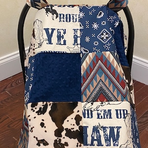 May include: A patchwork car seat cover with a cowboy theme. The cover features a blue bandana print, a brown and white cow print, and a red, white, and blue geometric pattern. The cover also has the text "Proud Ye Haw" and "No Em Up Law".