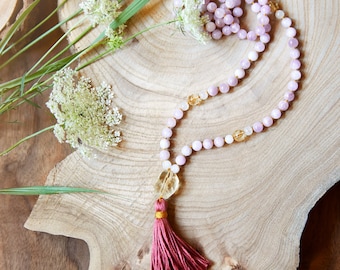 May include: A pink and yellow gemstone mala necklace with a pink tassel. The necklace is on a wooden surface with green plants.