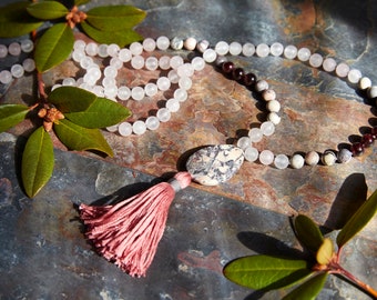 May include: A pink and white mala necklace with a large, oval-shaped stone bead. The necklace is made of natural stones and has a tassel at the end.