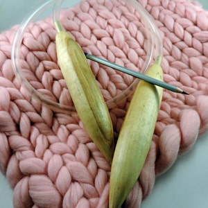 May include: Two wooden knitting needles with a clear plastic cable, resting on a pink knitted blanket.