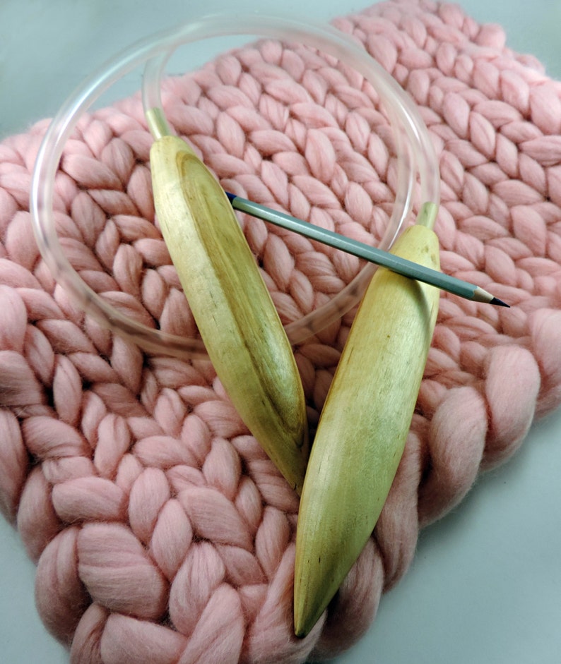May include: Two wooden knitting needles with a clear plastic cable, resting on a pink knitted blanket.