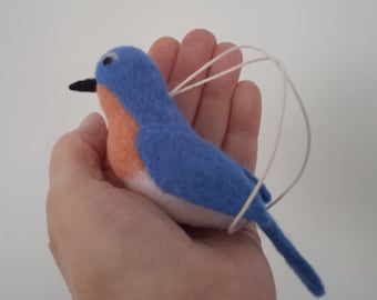 May include: A felt bluebird ornament with a white string for hanging. The bird has a light orange breast and a black beak.