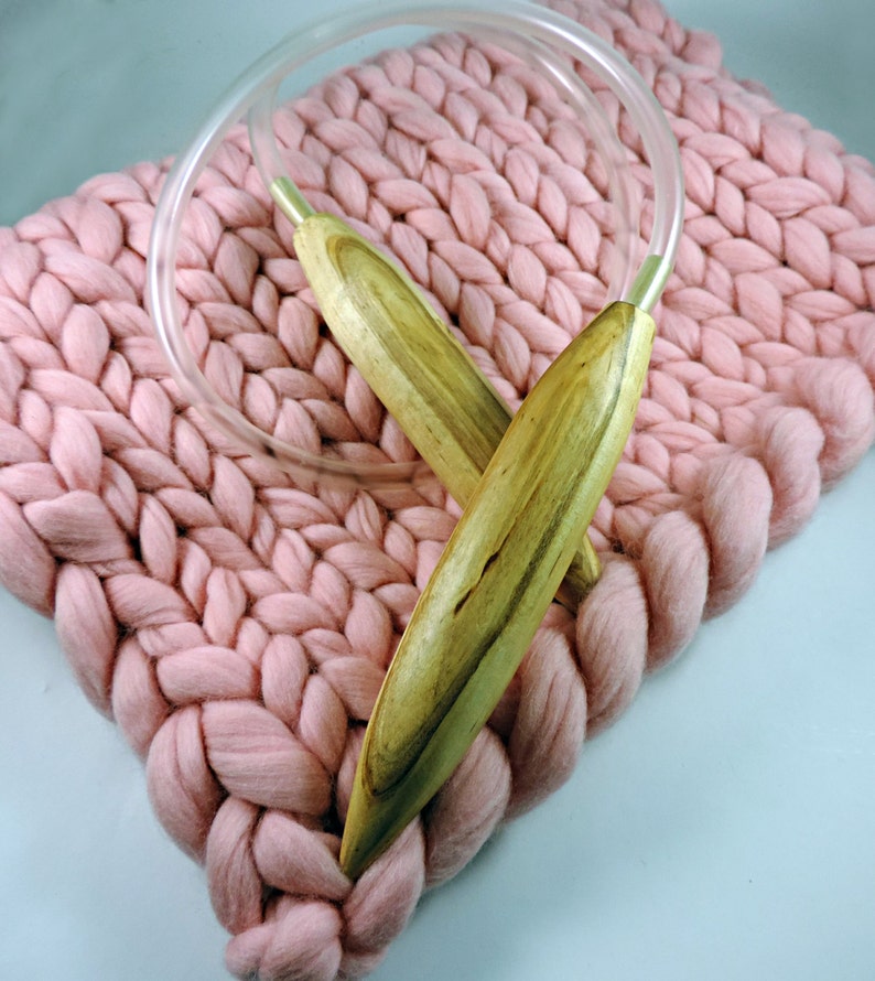 May include: A pair of wooden knitting needles with clear plastic tubing, resting on a pink knitted blanket.