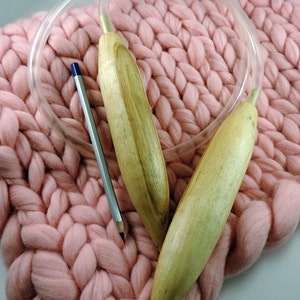 May include: Two wooden knitting needles with a clear plastic cable, resting on a pink knitted blanket.