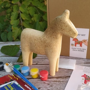 Può includere: Un kit artigianale di cavallo Dala con una statuina di cavallo in legno beige, vasetti di vernice, pennelli e pennarelli acrilici. La scatola recita "Paint Your Own Dala Horse". Il set include una guida.