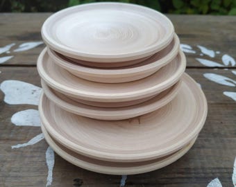 May include: A stack of six unfinished wooden plates. The plates are light brown with visible wood grain and a slightly raised rim. The plates are stacked on top of each other, with the largest plate at the bottom.