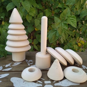 May include: A selection of unfinished wooden toys, featuring a tiered tree, a mallet, and diverse geometric forms. The natural wood grain is visible on each piece. The toys are arranged on a wooden surface against a backdrop of green foliage.
