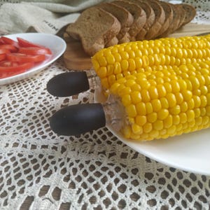 May include: Two yellow corn on the cob with black corn holders on a white plate. The corn is cooked and has a glossy sheen.