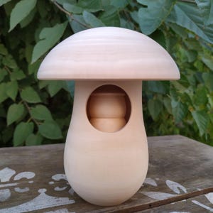 May include: A light-colored, wooden mushroom-shaped decorative item. The mushroom has a rounded cap and a stem with a circular opening. The item is made of unfinished wood, showcasing its natural grain. The background features green foliage.