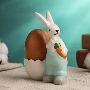 Könnte beinhalten: Eine weiße Keramik-Hasenfigur, die eine Karotte hält, mit einem braunen Ei in einem weißen Eierbecher.