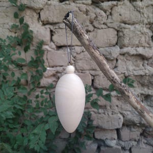 Pode incluir: Um ornamento de madeira branco em forma de lágrima, pendurado em um cordão cinza. O ornamento está suspenso de um galho contra um fundo de parede de pedra.
