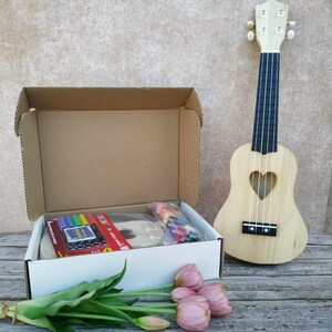 Könnte beinhalten: Eine Holzukulele mit herzförmigem Schallloch steht neben einer offenen Box mit Kunstbedarf. Die Box enthält Marker und eine Holzform. Rosa Tulpen befinden sich im Vordergrund.