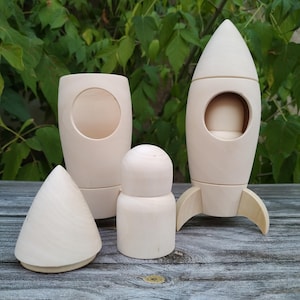 May include: A collection of unfinished wooden toys, including a rocket ship, a cylinder with a large circular opening, a small rounded figure, and a cone. The toys are made of light-colored wood and are unpainted.