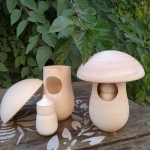 May include: A collection of unfinished wooden mushroom-shaped objects. The set includes a large mushroom with a rounded cap, a smaller mushroom-shaped container, and a small jar with a lid. The objects are displayed on a wooden surface.