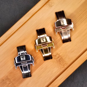 May include: Three watch clasps in silver, gold, and black, arranged on a wooden surface. Each clasp features a rectangular design with a central logo. The clasps are made of metal and have a polished finish, reflecting light.