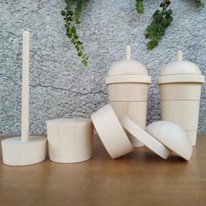 Puede incluir: Colección de juguetes de madera sin terminar, que incluye dos formas de taza con tapas, una base cilíndrica con una varilla vertical, un cilindro pequeño y dos piezas circulares. Los juguetes son de color beige claro y parecen hechos a mano.