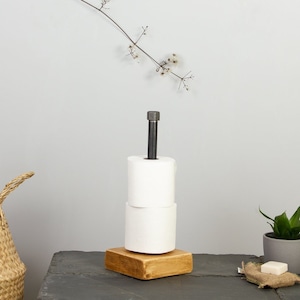 May include: A rustic toilet paper holder made of wood and metal. The holder has a square wooden base and a metal rod with a black finish. Two rolls of toilet paper are on the holder.