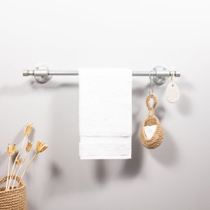 May include: A silver-grey metal towel rail with a white towel. A woven jute sponge and a pumice stone hang from the right side. A basket with dried flowers is in the lower left corner.