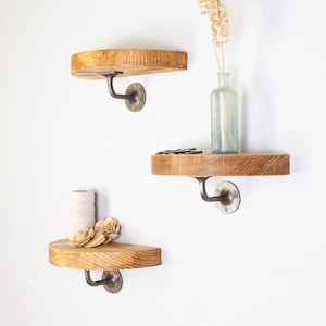 May include: Three wooden shelves with metal brackets mounted on a white wall. The shelves are half-circle shaped and have a natural wood finish. The brackets are a dark silver color.