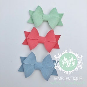 May include: Three felt hair bows in different colors: light blue, coral, and light green. The bows are arranged in a vertical stack, with the light blue bow at the bottom, the coral bow in the middle, and the light green bow at the top.