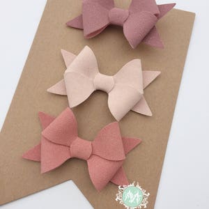 May include: Three felt hair bows in shades of pink, blush, and mauve. The bows are arranged in a triangular shape on a brown background. The text "MMBOWTIQUE custom. handmade" is visible at the bottom of the image.