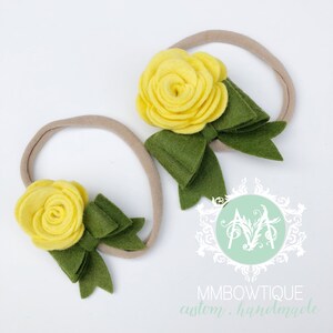 May include: Two nude elastic headbands with yellow felt flower and green felt bow accents. The headbands are handmade and the text "MMBOWTIQUE custom. handmade" is visible at the bottom of the image.