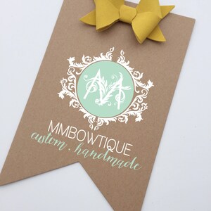 May include: A brown paper tag with a white logo featuring a stylized "MM" in a circle with a green background and a white floral design. The text "MMBOWTIQUE custom. handmade" is printed below the logo.