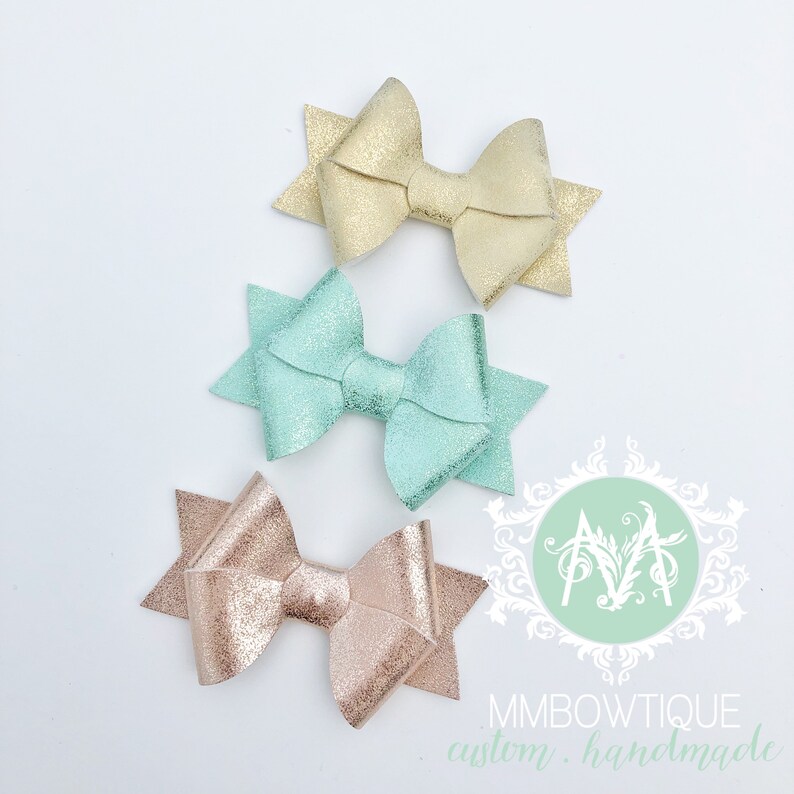 May include: Three sparkly hair bows in gold, mint green, and rose gold. The bows are handmade and have a starburst shape.