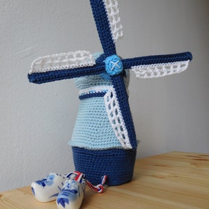 Puede incluir: Un molino de viento de crochet azul y blanco con detalles de encaje blanco. Dos zuecos de cerámica azul y blanco están en primer plano.