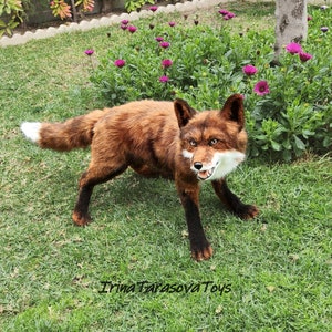 May include: A realistic plush fox with a red and brown fur coat, black legs, and a white tip on its tail. The fox is standing on green grass with purple flowers in the background. The text "Irina Tarasova Toys" is visible in the bottom right corner of the image.