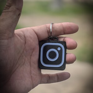 May include: Black square keychain with a light blue Instagram logo. The keychain is held in a hand, with a silver key ring attached. The background is blurred, suggesting an outdoor setting.