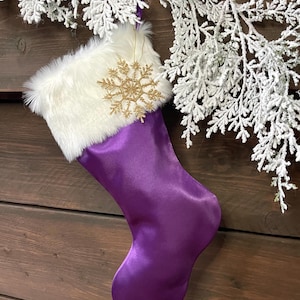 Pode incluir: Uma meia de Natal de cetim roxo com punho de pele sintética branca e um enfeite de floco de neve dourado. A meia está pendurada em uma superfície de madeira, com galhos brancos gelados ao fundo.