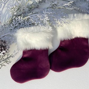 May include: Two burgundy velvet Christmas stockings with white faux fur trim. The stockings are displayed on a white surface with frosted greenery and a pinecone in the background. Festive holiday decor.