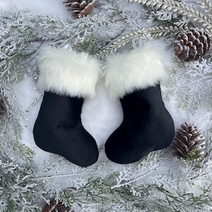 May include: Two black Christmas stockings with white faux fur trim. The stockings are displayed on a bed of white faux fur and surrounded by frosted greenery and pinecones, creating a festive holiday scene.