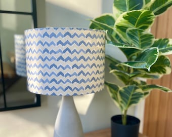 Könnte beinhalten: Eine graue Keramik-Tischlampe mit einem Lampenschirm mit blau-weißem Chevron-Muster. Die Lampe steht auf einem Holztisch neben einer Topfpflanze.