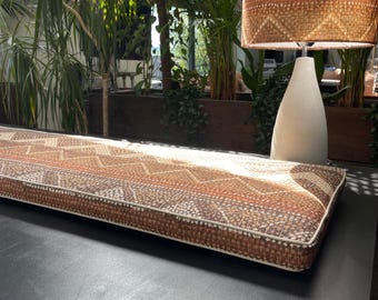May include: A long, rectangular bench cushion with a geometric pattern in shades of brown, beige, and orange. The cushion has a white border and is placed on a dark surface. A lamp with a woven shade and a white base is in the background.