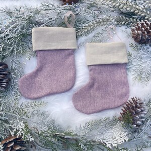May include: Two lavender Christmas stockings with a light beige cuff and a loop for hanging. The stockings are displayed on a bed of white faux fur, surrounded by frosted greenery and pinecones.
