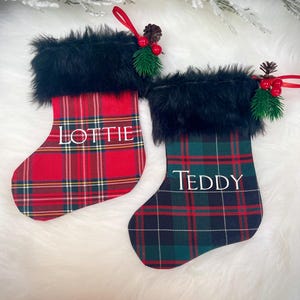 Peut inclure: Deux bas de Noël avec une bordure en fausse fourrure noire. L'un est à carreaux rouges avec le nom "Lottie" en blanc, l'autre à carreaux verts avec "Teddy". Chacun a une boucle de ruban rouge et des décorations de pommes de pin et de baies.
