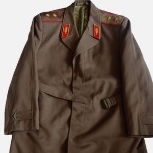 May include: A brown military-style coat with a belted waist and shoulder epaulets. The coat features red and gold accents on the collar and shoulders, with a double-breasted design and buttoned cuffs. The coat is a vintage style.