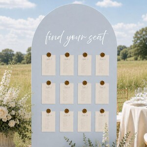 May include: A light blue arched seating chart sign with the text find your seat in white script. Twelve cream-colored cards are attached to the board with gold wax seals, arranged in three columns of four, displaying guest names and table numbers.