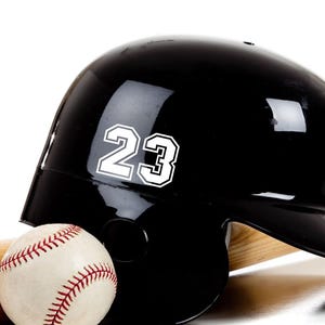 May include: A black baseball helmet with a white number 23 on the side. A baseball and a wooden bat are in the background.