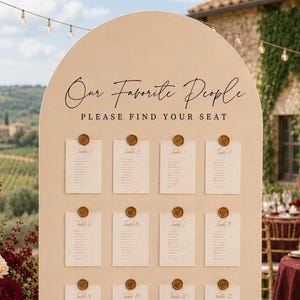May include: A large, arched, beige seating chart sign with the text "Our Favorite People" and "PLEASE FIND YOUR SEAT" in black. Twelve white rectangular cards are attached to the board with gold wax seals, each listing table numbers.