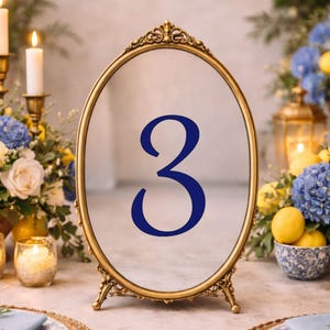 May include: An oval table number sign featuring a gold-toned metal frame with ornate detailing at the top and base. The center of the sign displays the number 3 in a bold, dark blue serif font, positioned upright on a flat surface.