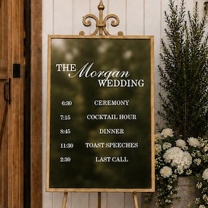 May include: A rectangular dark green wedding schedule sign with white text reading THE MORGAN WEDDING, CEREMONY, COCKTAIL HOUR, DINNER, TOAST SPEECHES, and LAST CALL, displayed on a gold metal easel with a decorative fleur-de-lis top finial.